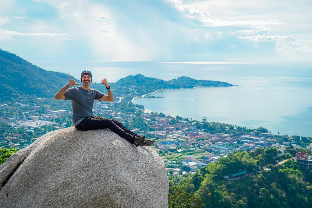A man sitting on a massive granite boulder, posing for a photo with a panoramic view of Koh Samui, Thailand behind him. The background shows lush green hills, the Lamai valley, and the ocean under a bright sky.