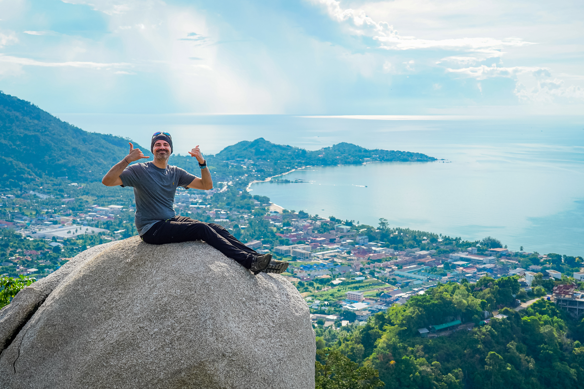 A man sitting on a massive granite boulder, posing for a photo with a panoramic view of Koh Samui, Thailand behind him. The background shows lush green hills, the Lamai valley, and the ocean under a bright sky.